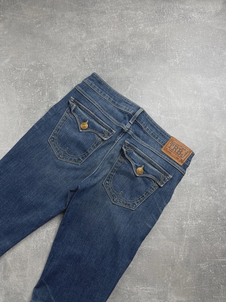 May include: Blue denim jeans featuring two back pockets with button-down flaps, decorative stitching, and a brown leather patch on the waistband embossed with the text TRBJ. The jeans have a classic medium wash finish and visible belt loops.