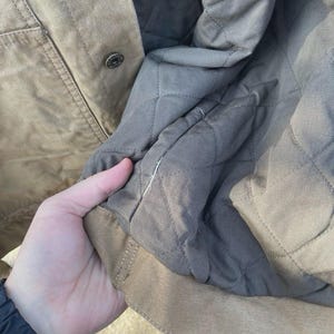 Puede incluir: Una chaqueta de color tostado con un forro acolchado gris oscuro. El exterior es de color tostado s&oacute;lido con un bot&oacute;n de presi&oacute;n met&aacute;lico visible. El interior presenta un patr&oacute;n de costuras en forma de diamante en tela gris oscuro.