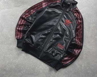 May include: A black track jacket with a shiny finish, featuring a high collar and a full-length front zip. The sleeves are adorned with three red stripes. A red Adidas trefoil logo is on the chest, with an Adidas Chile 62 patch below it.