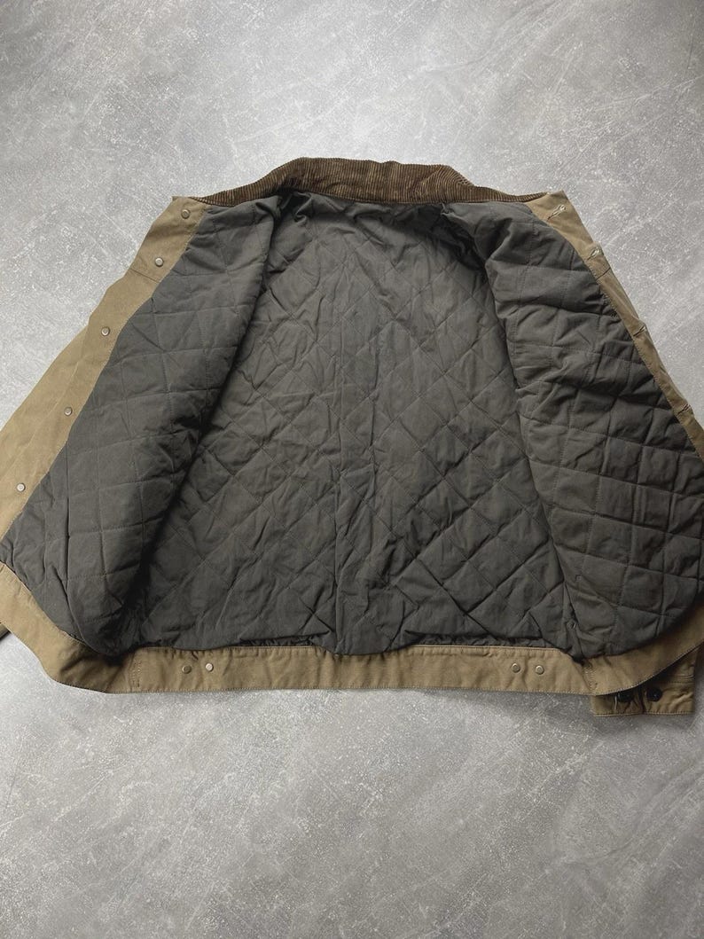 Puede incluir: Una chaqueta de color tostado con cuello de pana marr&oacute;n oscuro y forro acolchado gris oscuro. La chaqueta presenta una abertura frontal con botones de presi&oacute;n met&aacute;licos a lo largo de la tapeta y los pu&ntilde;os, con costuras visibles.