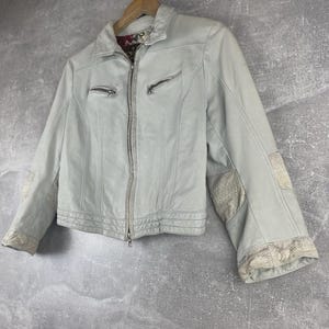 Peut inclure: Veste en cuir bleu clair avec fermeture &eacute;clair et deux poches zipp&eacute;es. La veste pr&eacute;sente des coutures d&eacute;coratives et des empi&egrave;cements &agrave; motifs sur les manches. Elle est suspendue &agrave; un cintre en bois sur fond gris.