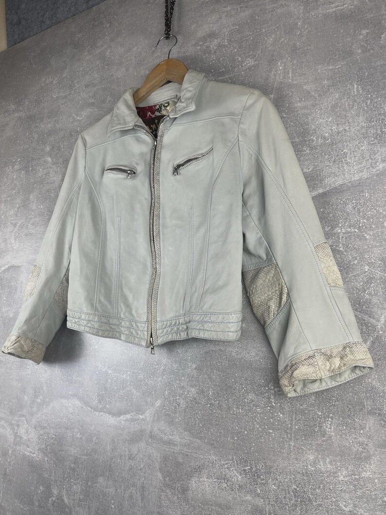 Peut inclure: Veste en cuir bleu clair avec fermeture &eacute;clair et deux poches zipp&eacute;es. La veste pr&eacute;sente des coutures d&eacute;coratives et des empi&egrave;cements &agrave; motifs sur les manches. Elle est suspendue &agrave; un cintre en bois sur fond gris.