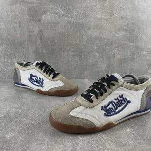 May include: A pair of low-top sneakers featuring white leather panels, beige suede overlays, and black laces. The side of each shoe displays the blue embroidered text Von Dutch. The design includes a brown rubber sole and blue heel accents.