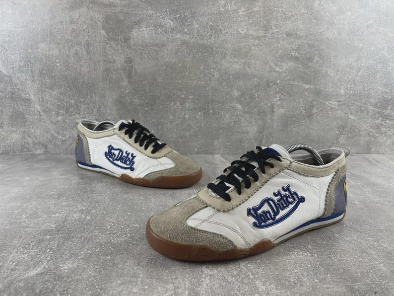 May include: A pair of low-top sneakers featuring white leather panels, beige suede overlays, and black laces. The side of each shoe displays the blue embroidered text Von Dutch. The design includes a brown rubber sole and blue heel accents.
