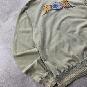 May include: Sage green hoodie featuring a winged eye graphic on the back. The hoodie has a drawstring hood, ribbed cuffs, and a striped hem. The graphic shows yellow wings and a blue and white eye.