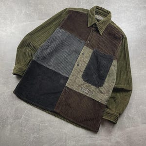 Peut inclure: Chemise en velours côtelé à manches longues, de style patchwork. Elle présente des panneaux de velours côtelé vert olive, noir, gris et marron. Boutonnage, col et une poche de poitrine.