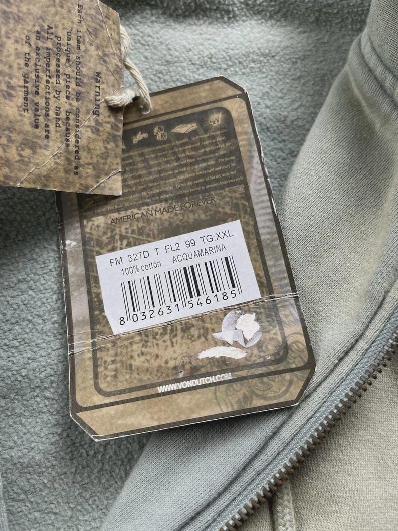 May include: A light green hoodie with a zip closure, viewed close up. A tag is attached, showing the text "AMERICAN MADE FOREVER", product details, and a barcode. The hoodie is made of 100% cotton.