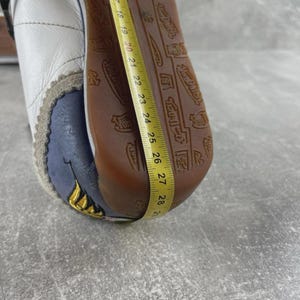May include: A white leather sneaker with a brown rubber sole featuring a repeating Von Dutch logo pattern. A yellow measuring tape is placed along the sole, showing a length measurement extending from 14 to 29 centimeters on the metric side.