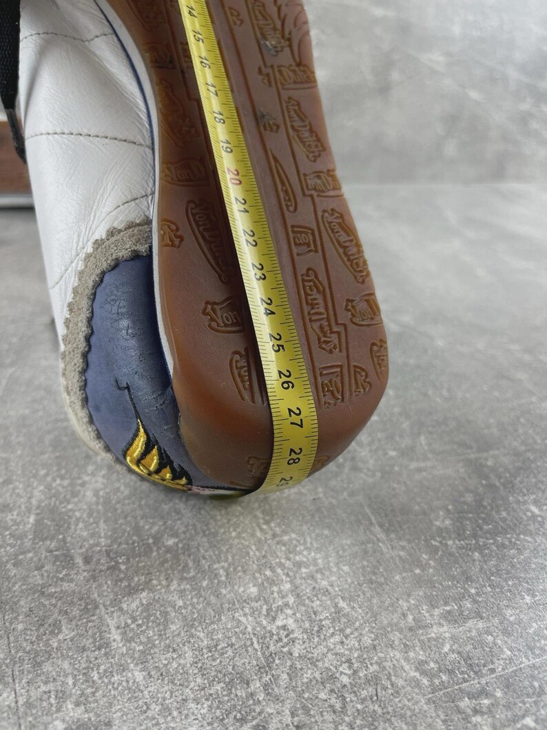 May include: A white leather sneaker with a brown rubber sole featuring a repeating Von Dutch logo pattern. A yellow measuring tape is placed along the sole, showing a length measurement extending from 14 to 29 centimeters on the metric side.