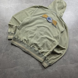 May include: A sage green hoodie with a drawstring hood and ribbed cuffs and hem. The back features a graphic of a blue and white eye with yellow wings. The hoodie is made of a soft, comfortable material.