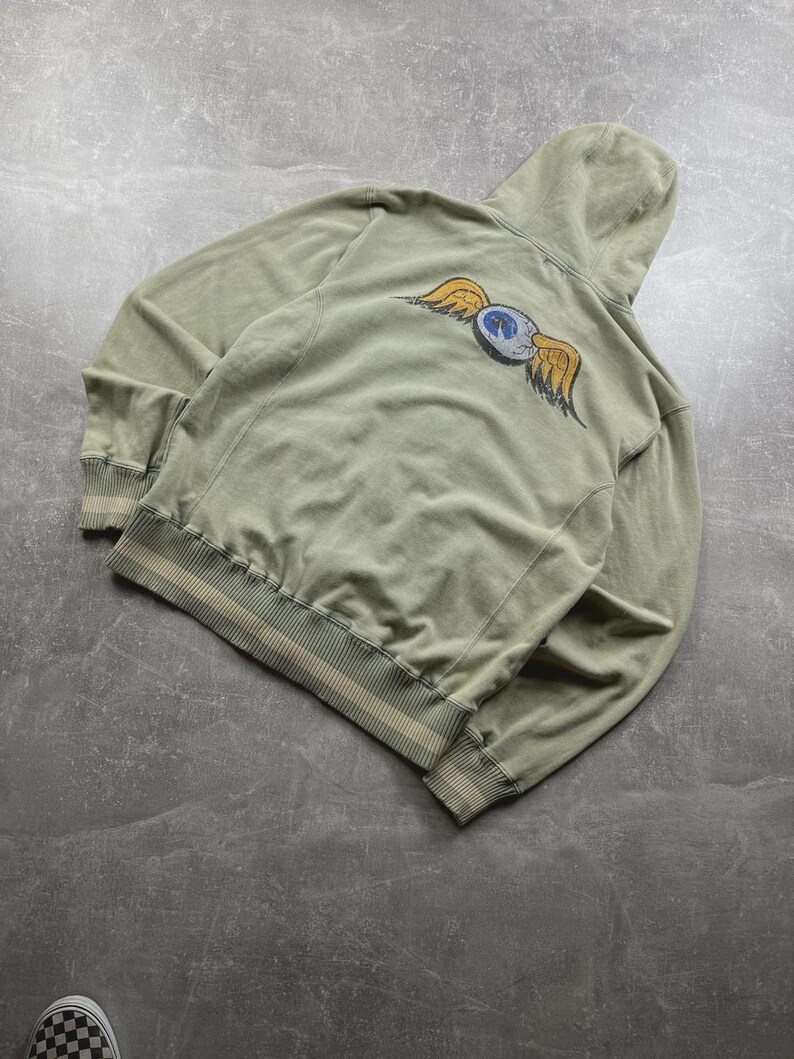 May include: A sage green hoodie with a drawstring hood and ribbed cuffs and hem. The back features a graphic of a blue and white eye with yellow wings. The hoodie is made of a soft, comfortable material.