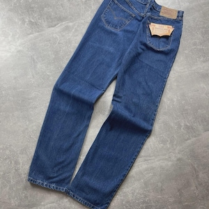 May include: A pair of dark blue denim jeans featuring a high-waisted cut, two back pockets with stitched detailing, a tan leather-like brand patch on the waistband, and a small orange brand tag attached to the right back pocket with visible text.