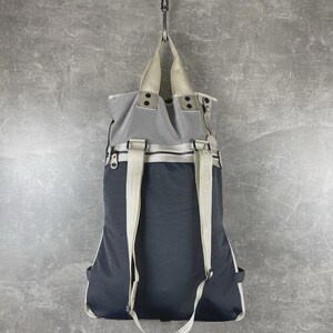 May include: A two-tone backpack featuring a dark navy blue lower section and a light gray mesh upper section. It includes beige shoulder straps, top carry handles, and a horizontal zipper closure separating the two distinct fabric panels.