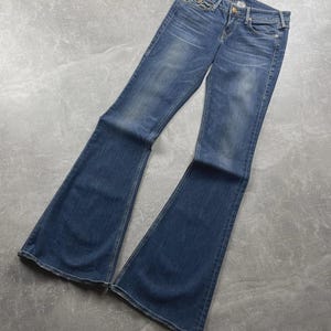May include: Blue denim jeans featuring a flared leg silhouette, a mid-rise waist, a button and zipper fly closure, and classic five-pocket styling. The fabric displays a faded wash effect on the thighs and knees with visible stitching details.