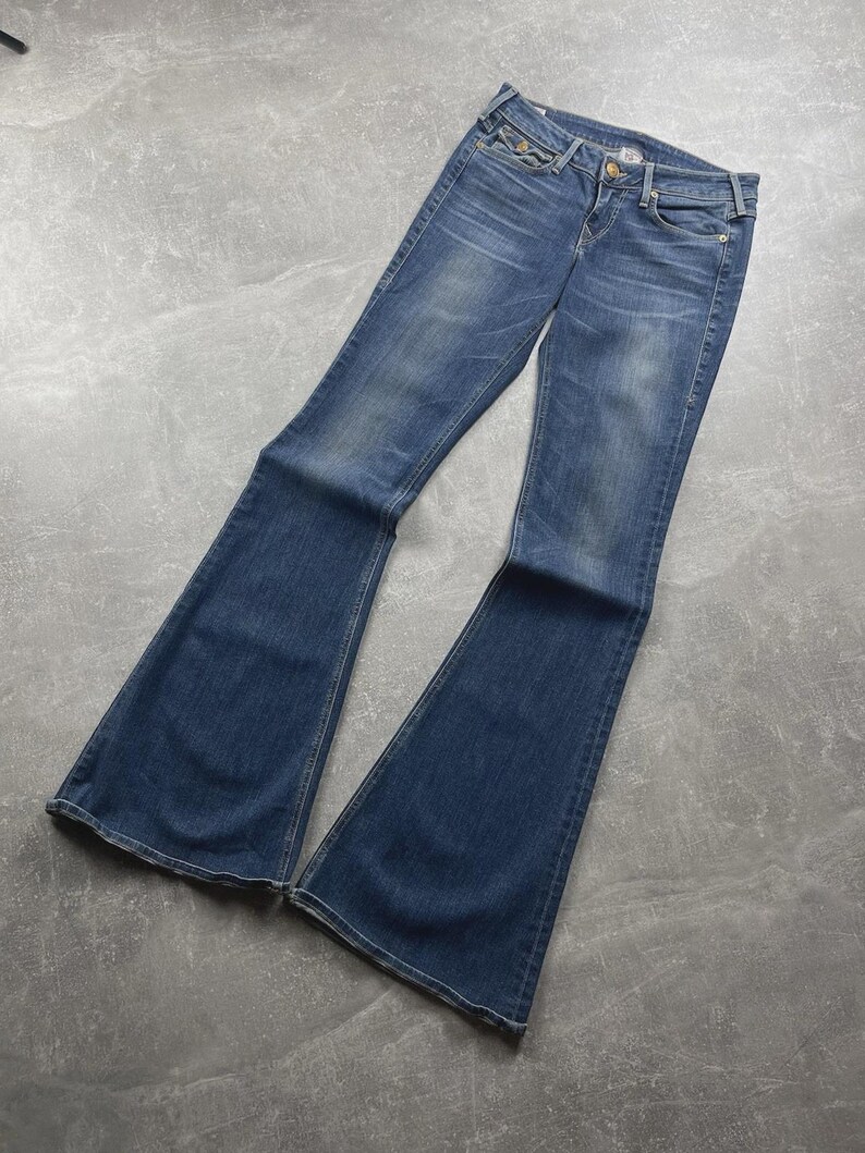 May include: Blue denim jeans featuring a flared leg silhouette, a mid-rise waist, a button and zipper fly closure, and classic five-pocket styling. The fabric displays a faded wash effect on the thighs and knees with visible stitching details.