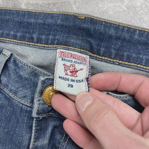 May include: Blue denim jeans featuring a white interior tag with the text True Religion Brand Jeans, a red graphic of a person playing a guitar, the text Made in USA, and the number 29. The jeans have gold-toned metal hardware and yellow stitching.