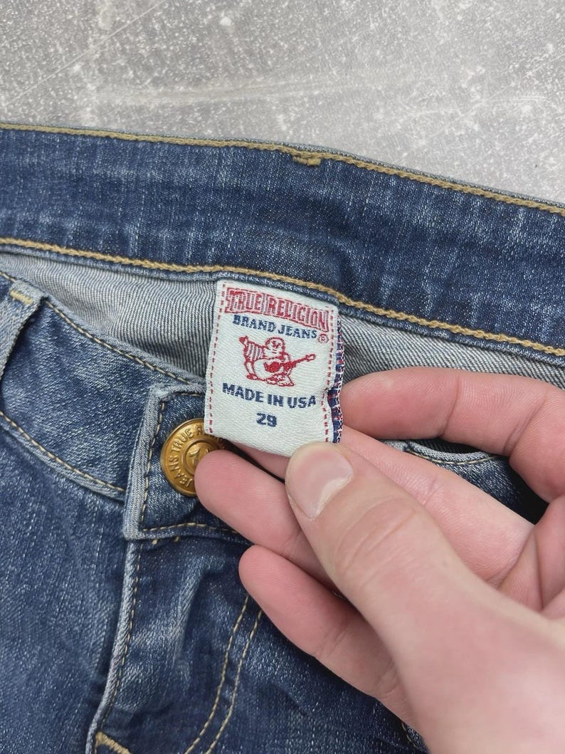 May include: Blue denim jeans featuring a white interior tag with the text True Religion Brand Jeans, a red graphic of a person playing a guitar, the text Made in USA, and the number 29. The jeans have gold-toned metal hardware and yellow stitching.