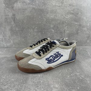 May include: A pair of white and beige sneakers featuring black laces and the blue Von Dutch logo embroidered on the side. The shoes have a brown rubber sole, suede overlays on the toe and eyelets, and a blue heel panel with a yellow circular emblem.