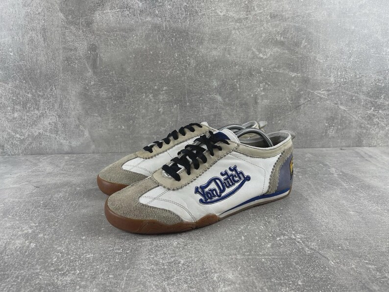May include: A pair of white and beige sneakers featuring black laces and the blue Von Dutch logo embroidered on the side. The shoes have a brown rubber sole, suede overlays on the toe and eyelets, and a blue heel panel with a yellow circular emblem.