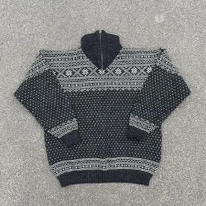 May include: A charcoal gray and white knit sweater featuring a quarter-zip high collar. The garment displays a detailed Nordic-inspired geometric pattern across the chest and sleeves, complemented by a subtle dotted texture throughout the body.