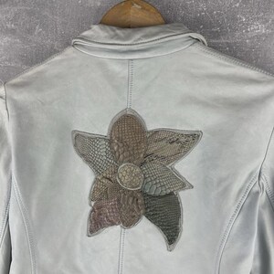 Peut inclure: Veste en cuir bleu clair orn&eacute;e d'une fleur sur le dos. La fleur est compos&eacute;e de mati&egrave;res textur&eacute;es dans des tons marron, vert et gris. La veste est suspendue &agrave; un cintre en bois.