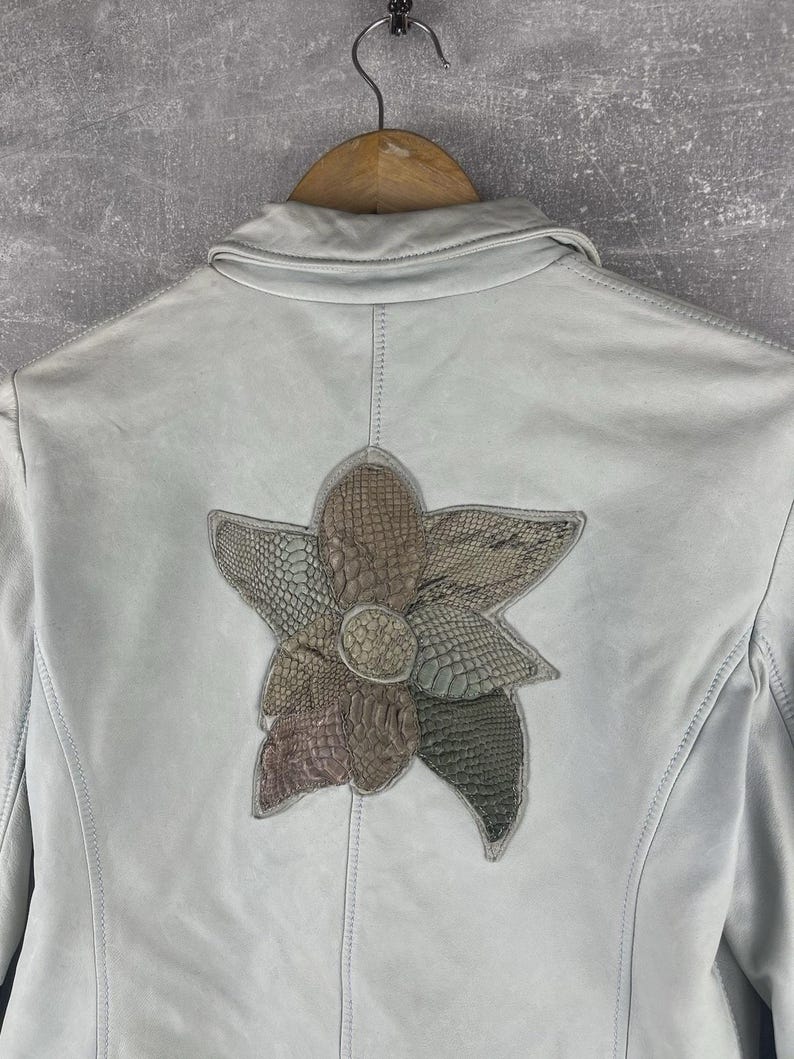 Peut inclure: Veste en cuir bleu clair orn&eacute;e d'une fleur sur le dos. La fleur est compos&eacute;e de mati&egrave;res textur&eacute;es dans des tons marron, vert et gris. La veste est suspendue &agrave; un cintre en bois.