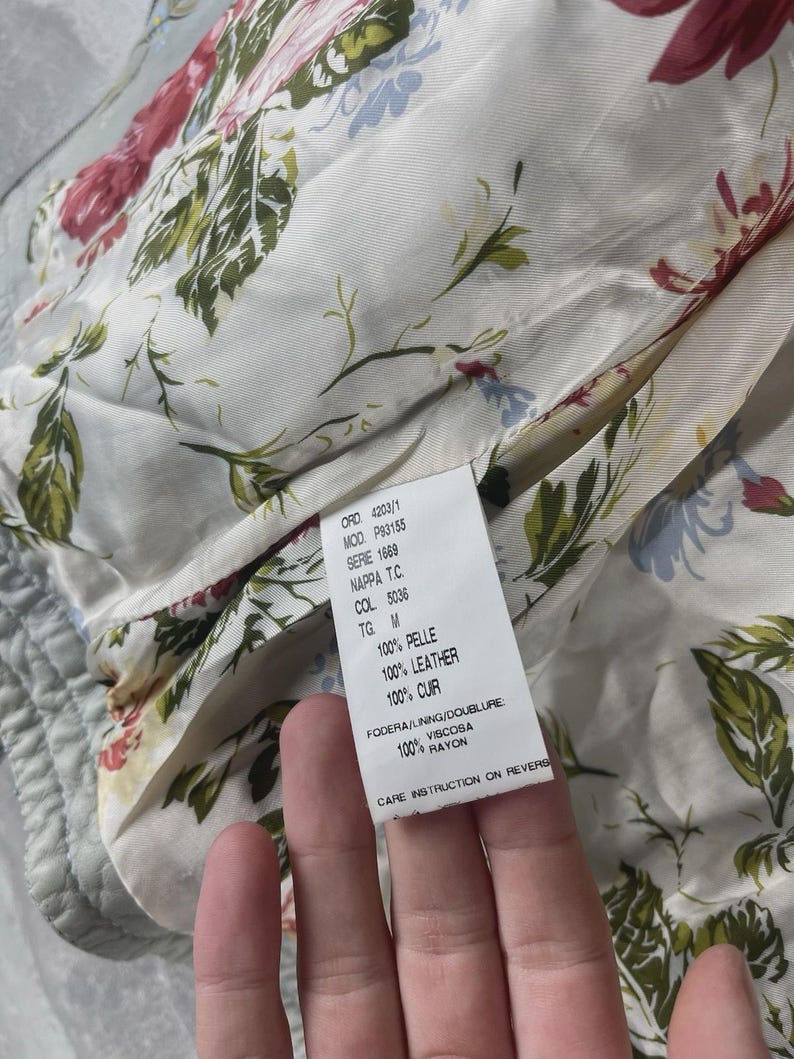 Peut inclure: Gros plan sur la doublure int&eacute;rieure d'un v&ecirc;tement, avec un motif floral dans les tons de rouge, vert et bleu sur fond cr&egrave;me. Une &eacute;tiquette avec du texte en plusieurs langues est visible, indiquant la composition du mat&eacute;riau et les instructions d'entretien.