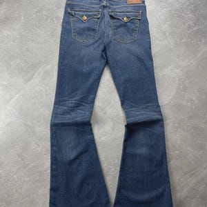 May include: Dark blue denim flare jeans featuring a mid-rise waist, two back pockets with button-down flaps, and a tan leather brand patch on the waistband. The jeans exhibit subtle whiskering and fading patterns along the thighs and knees.
