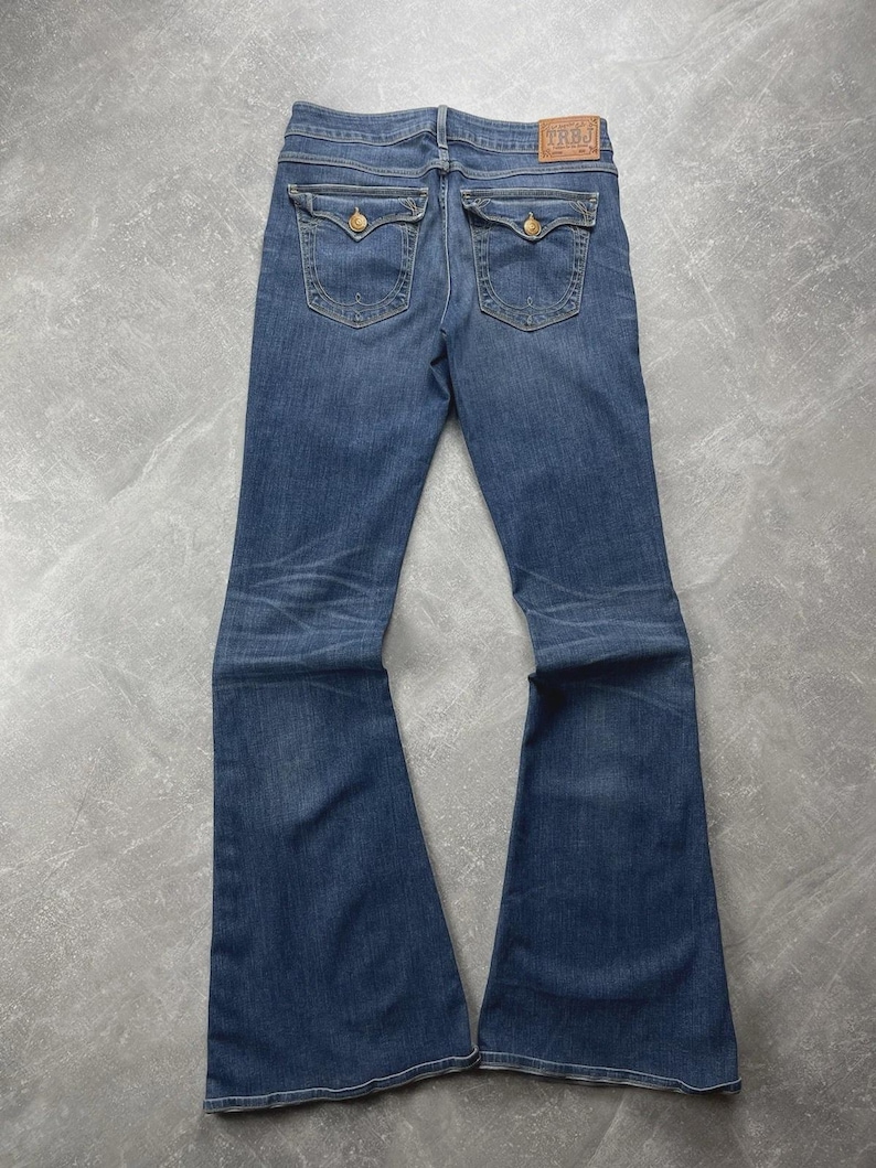 May include: Dark blue denim flare jeans featuring a mid-rise waist, two back pockets with button-down flaps, and a tan leather brand patch on the waistband. The jeans exhibit subtle whiskering and fading patterns along the thighs and knees.