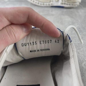 May include: White leather shoe with blue trim and white laces. The shoe's interior displays the text "DU1135 E7007 42" and "MADE IN ROMANIA". The sole is white with a blue accent.
