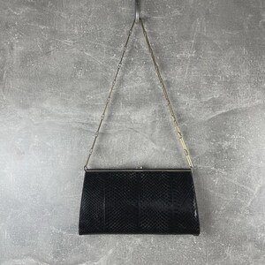 以下が含まれることがあります： 黒色の長方形クラッチバッグ。蛇革のような質感の模様が施されています。シルバーカラーの金属フレームと繊細なチェーンストラップが付いています。グレーの背景に吊り下げられています。
