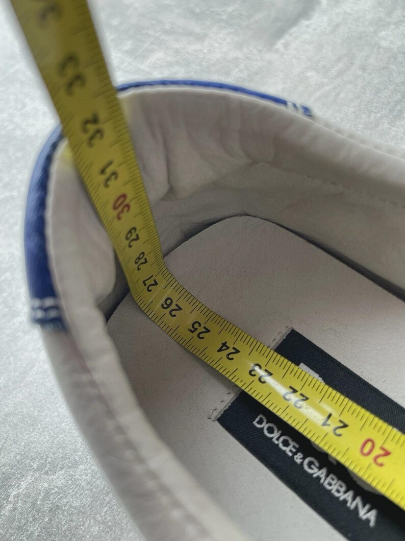 May include: White shoe with blue trim, measured with a yellow tape measure. The tape measure shows a length of approximately 23 cm. The shoe has a black label that reads "DOLCE & GABBANA".