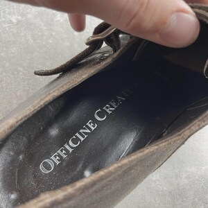 May include: Close-up of a brown leather shoe with a buckle closure. The shoe's interior features the text "OFFICINE CREATIVE" in silver lettering. The leather has a distressed appearance.