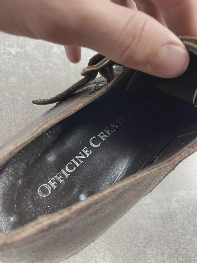 May include: Close-up of a brown leather shoe with a buckle closure. The shoe's interior features the text "OFFICINE CREATIVE" in silver lettering. The leather has a distressed appearance.