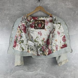 Peut inclure: Veste bleu clair avec une doublure int&eacute;rieure &agrave; motifs floraux. La veste a une fermeture &eacute;clair sur le devant et des d&eacute;tails en dentelle sur les poignets. La doublure int&eacute;rieure pr&eacute;sente un motif de roses rouges et roses sur fond blanc. L'&eacute;tiquette indique "Santa Croce Original".