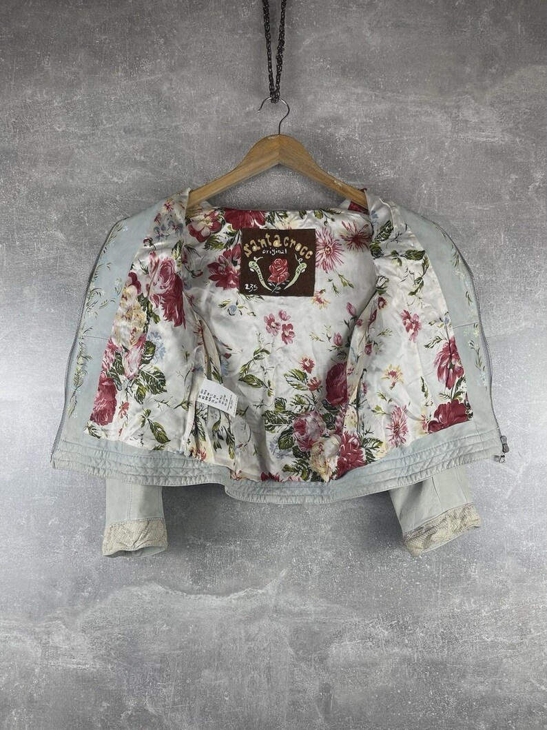 Peut inclure: Veste bleu clair avec une doublure int&eacute;rieure &agrave; motifs floraux. La veste a une fermeture &eacute;clair sur le devant et des d&eacute;tails en dentelle sur les poignets. La doublure int&eacute;rieure pr&eacute;sente un motif de roses rouges et roses sur fond blanc. L'&eacute;tiquette indique "Santa Croce Original".