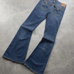 May include: Medium-wash blue denim flare jeans featuring a high-rise waist, two back pockets with button-down flaps, and a tan leather brand patch on the rear waistband. The jeans exhibit subtle whiskering and fading along the thighs and knees.