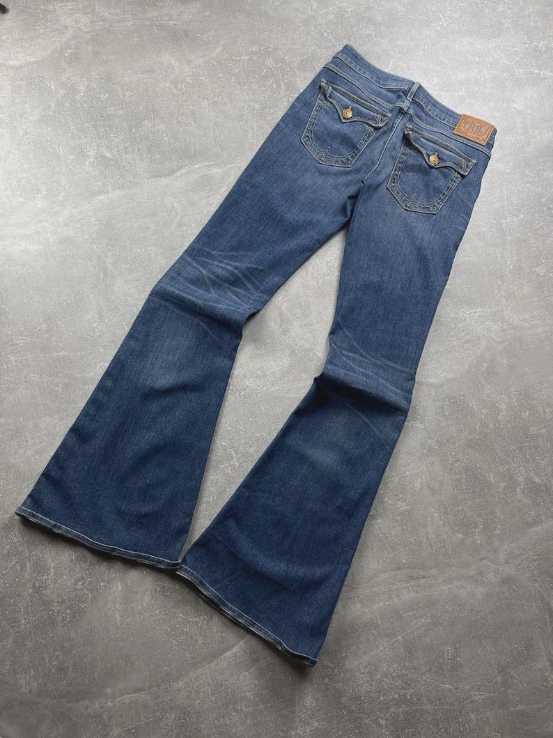 May include: Medium-wash blue denim flare jeans featuring a high-rise waist, two back pockets with button-down flaps, and a tan leather brand patch on the rear waistband. The jeans exhibit subtle whiskering and fading along the thighs and knees.