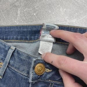 May include: Blue denim jeans featuring a gold-toned metal button embossed with the True Religion logo. A white fabric care tag is attached to the interior waistband, displaying text including RN#112790, CA#30427, and style number WELP73M47.