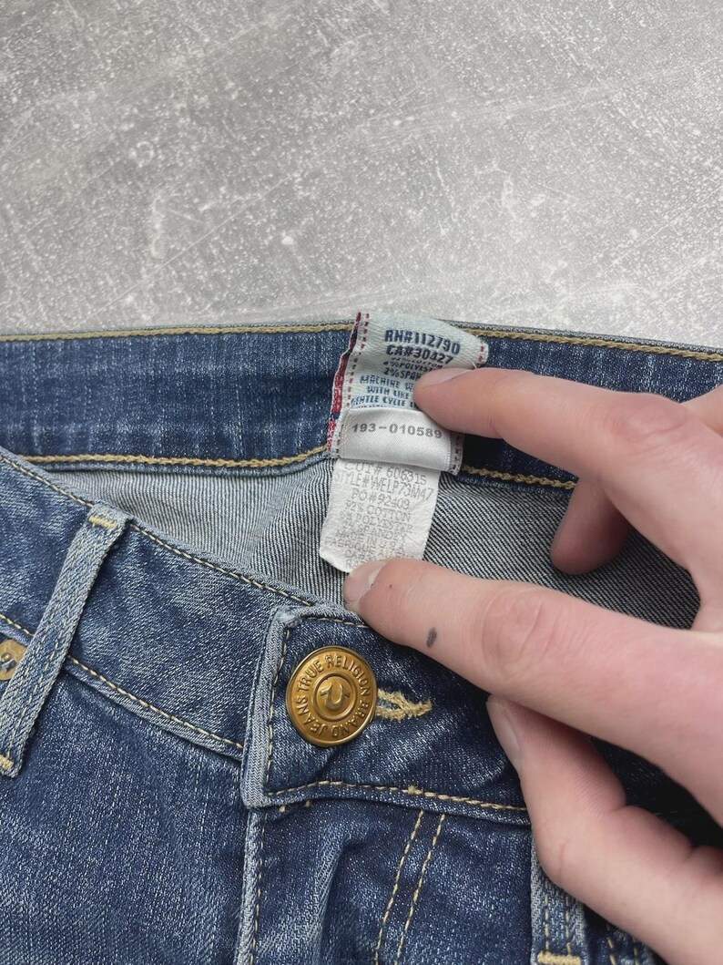 May include: Blue denim jeans featuring a gold-toned metal button embossed with the True Religion logo. A white fabric care tag is attached to the interior waistband, displaying text including RN#112790, CA#30427, and style number WELP73M47.