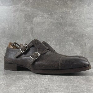 May include: A pair of brown leather double monk strap shoes. The shoes have silver buckles and a textured finish. The shoes are on a gray surface.