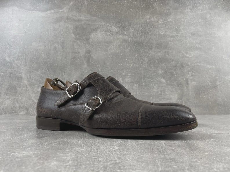 May include: A pair of brown leather double monk strap shoes. The shoes have silver buckles and a textured finish. The shoes are on a gray surface.
