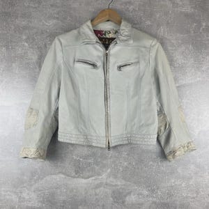 Puede incluir: Chaqueta de cuero gris claro con cremallera, dos bolsillos con cremallera y costuras decorativas. La chaqueta tiene detalles de tela estampada en las mangas y un forro con estampado floral. Colgada de una percha de madera.