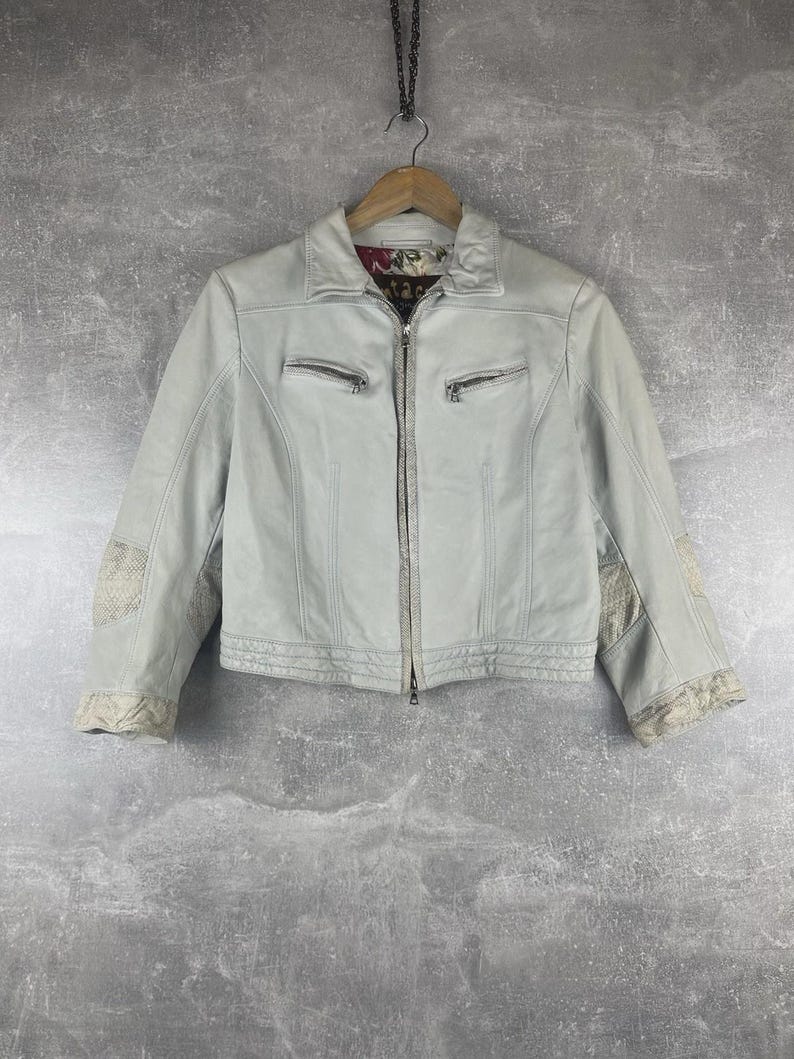 Peut inclure: Veste en cuir gris clair avec fermeture &eacute;clair, deux poches zipp&eacute;es et coutures d&eacute;coratives. La veste pr&eacute;sente des empi&egrave;cements en tissu &agrave; motifs sur les manches et une doublure &agrave; motifs floraux. Suspendue &agrave; un cintre en bois.