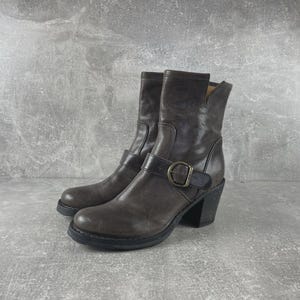 May include: A pair of dark brown leather ankle boots featuring a rounded toe, a stacked block heel, and a decorative strap across the midfoot secured with a metallic buckle. The boots have a mid-calf height with a notched detail at the top collar.