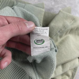 May include: A close-up of a pale green sweatshirt, showing a ribbed cuff and a label. The label reads "SPACE 2000" and "PAESE DI ORIGINE CINA". The fabric of the sweatshirt appears soft and textured.