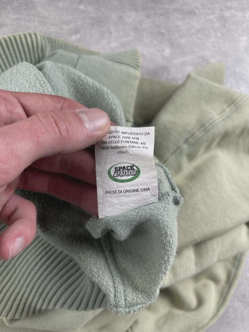 May include: A close-up of a pale green sweatshirt, showing a ribbed cuff and a label. The label reads "SPACE 2000" and "PAESE DI ORIGINE CINA". The fabric of the sweatshirt appears soft and textured.