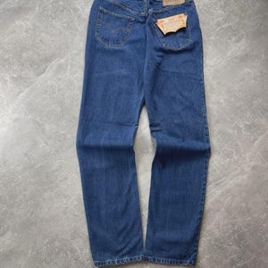 May include: A pair of blue denim jeans featuring a classic five-pocket design, a tan leather patch on the back waistband, and a rectangular paper tag attached to the right back pocket. The jeans have a medium wash and visible stitching details.