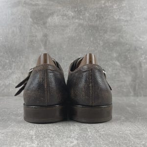 May include: A pair of brown leather shoes with a distressed finish. The shoes have a buckle closure and a low heel. The image shows the back of the shoes, with the heels and the buckle straps visible. The shoes appear to be well-worn.