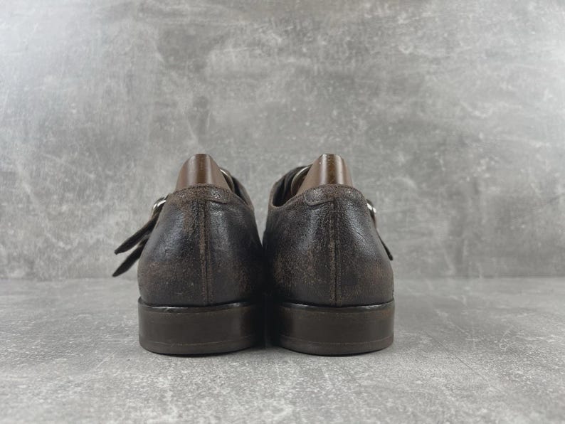 May include: A pair of brown leather shoes with a distressed finish. The shoes have a buckle closure and a low heel. The image shows the back of the shoes, with the heels and the buckle straps visible. The shoes appear to be well-worn.